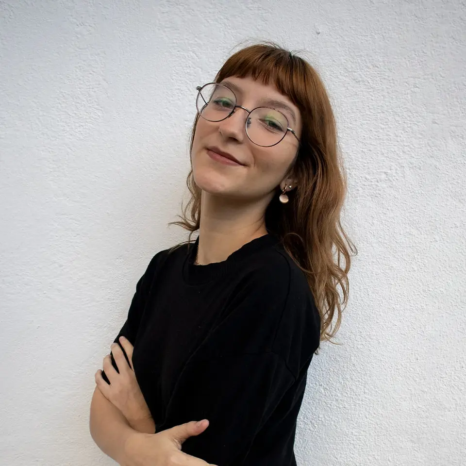 Retrato de Mylena Leite, de óculos e camiseta preta, sorrindo com a cabeça inclinada diante de uma parede branca.